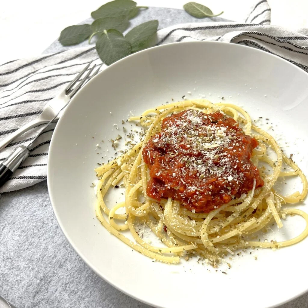 Pasta Mit Frischer Tomatensauce auf Teller mit Parmesan auf grauem Untergrund mit Besteck und weissem Stofftuch und Blättern im Hintergrund