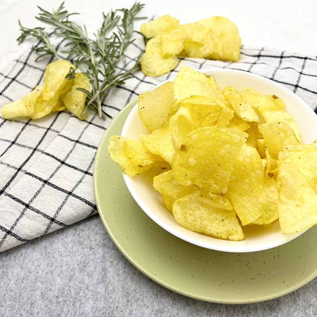 Kartoffelchips selber machen Pfanne gelb in weissem teller auf grünem teller auf grauem weissen stoff untergrund mit grünem thymian im hintergrund