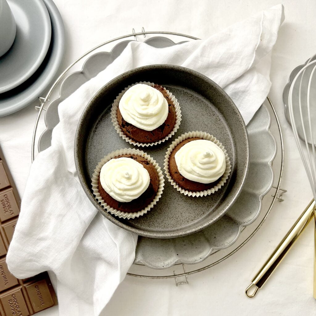 low carb cupcakes ohne zucker mit weisser sahne und braunem teig auf grauem teller auf weissemuntergrund neben grauem teller