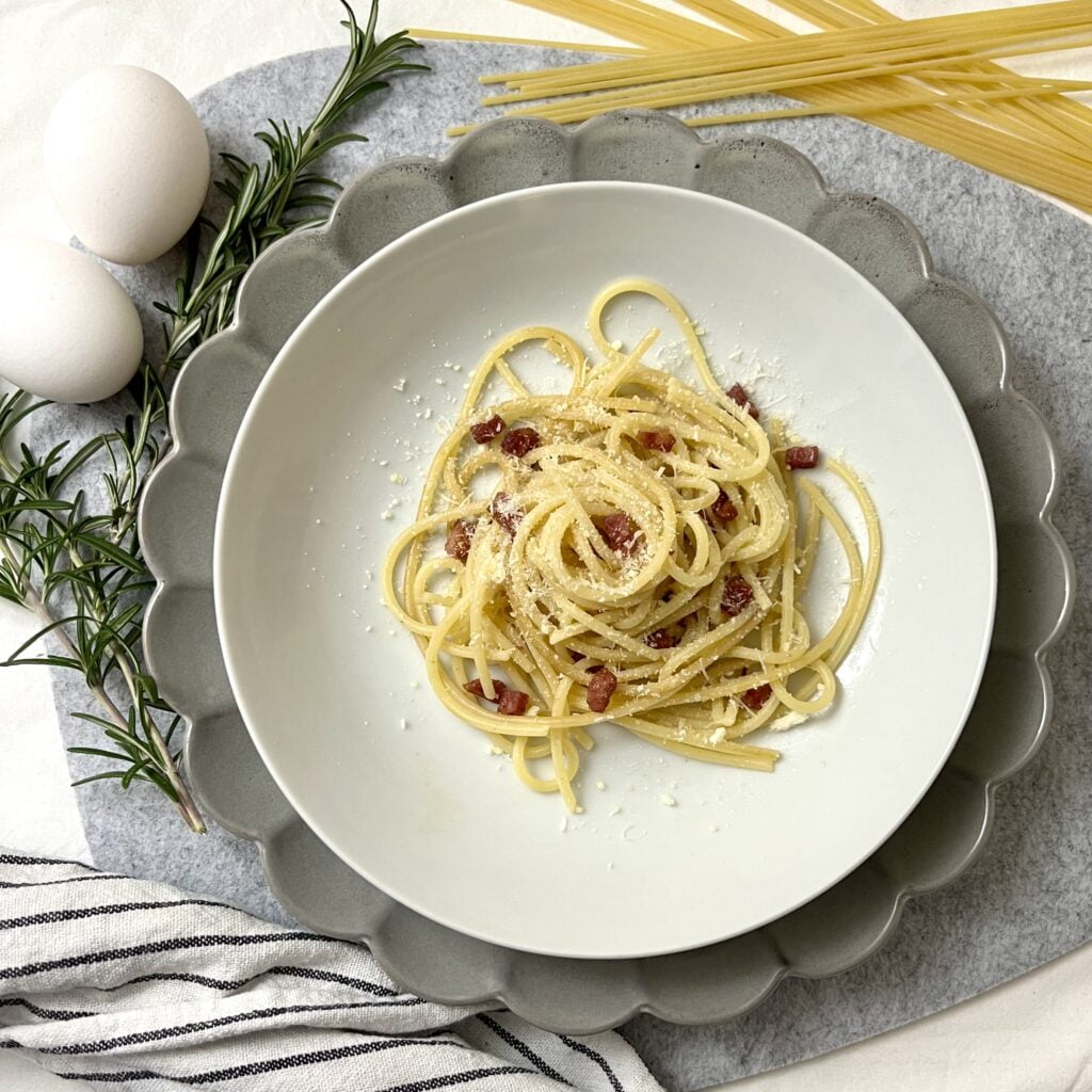 Traditionelle Spaghetti Carbonara Auf weissem Teller auf grauem teller auf grauem untergrund mit weissem stofftuch und grünen kräutern