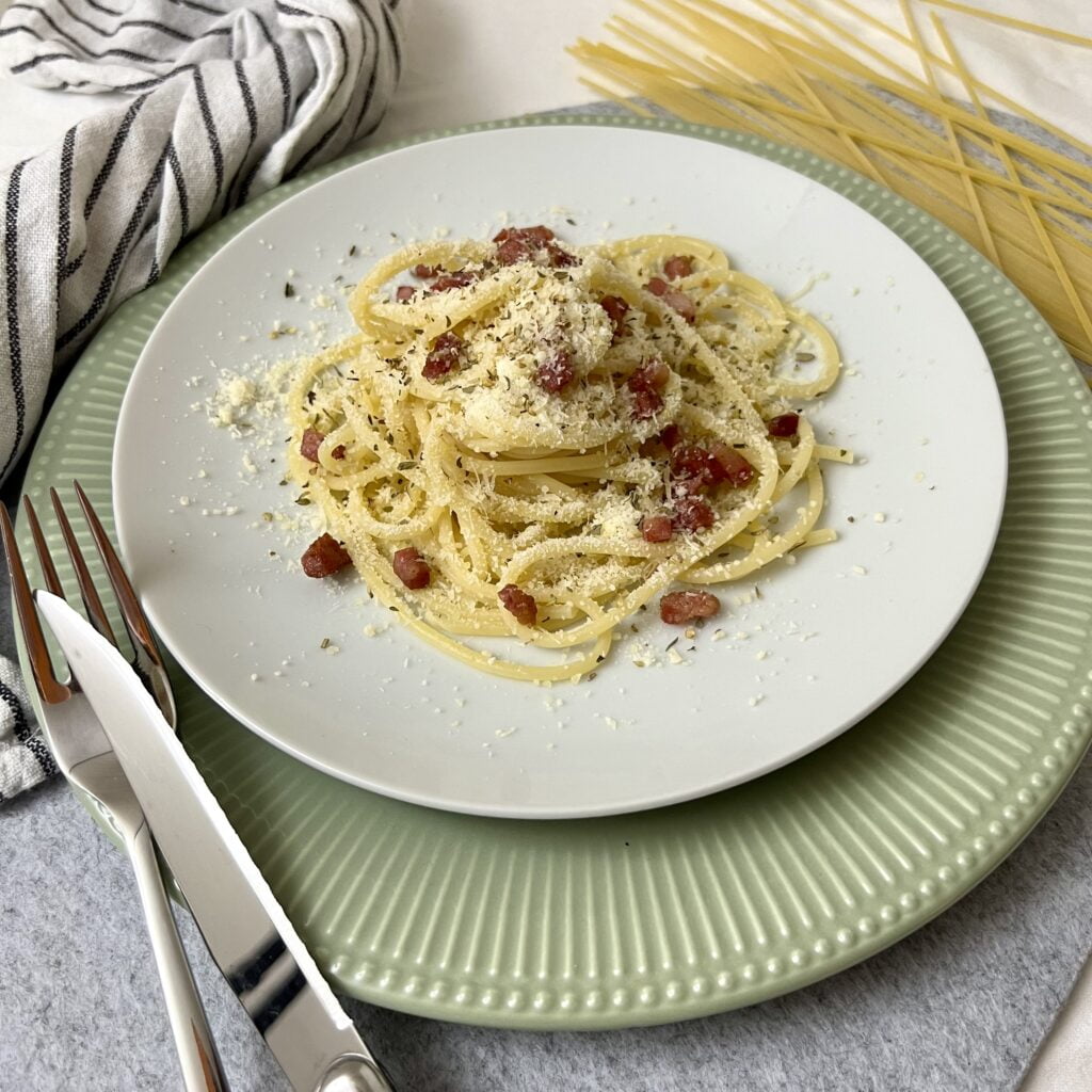 spaghetti carbonara mit speck und parmesan auf weissem teller auf grünem teller auf grauem untergrund mit besteck im vordergrund