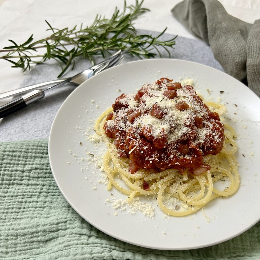 Bolognese Sauce rot auf gelben spaghetti mit weissem parmesan auf grünem tuch mit besteck und grünem rosmarin im hintergrund