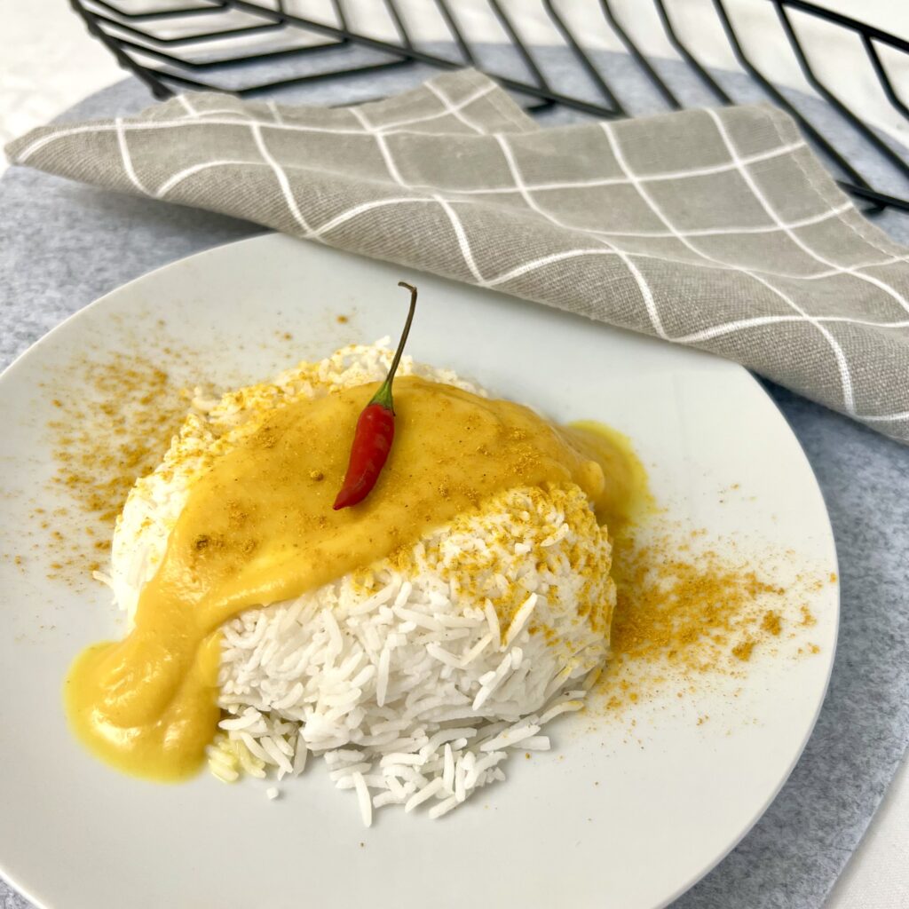 Gesundes Curry gelb auf weissem reis auf weissem teller neben grauem stofftuch mit roter chilli oben drauf