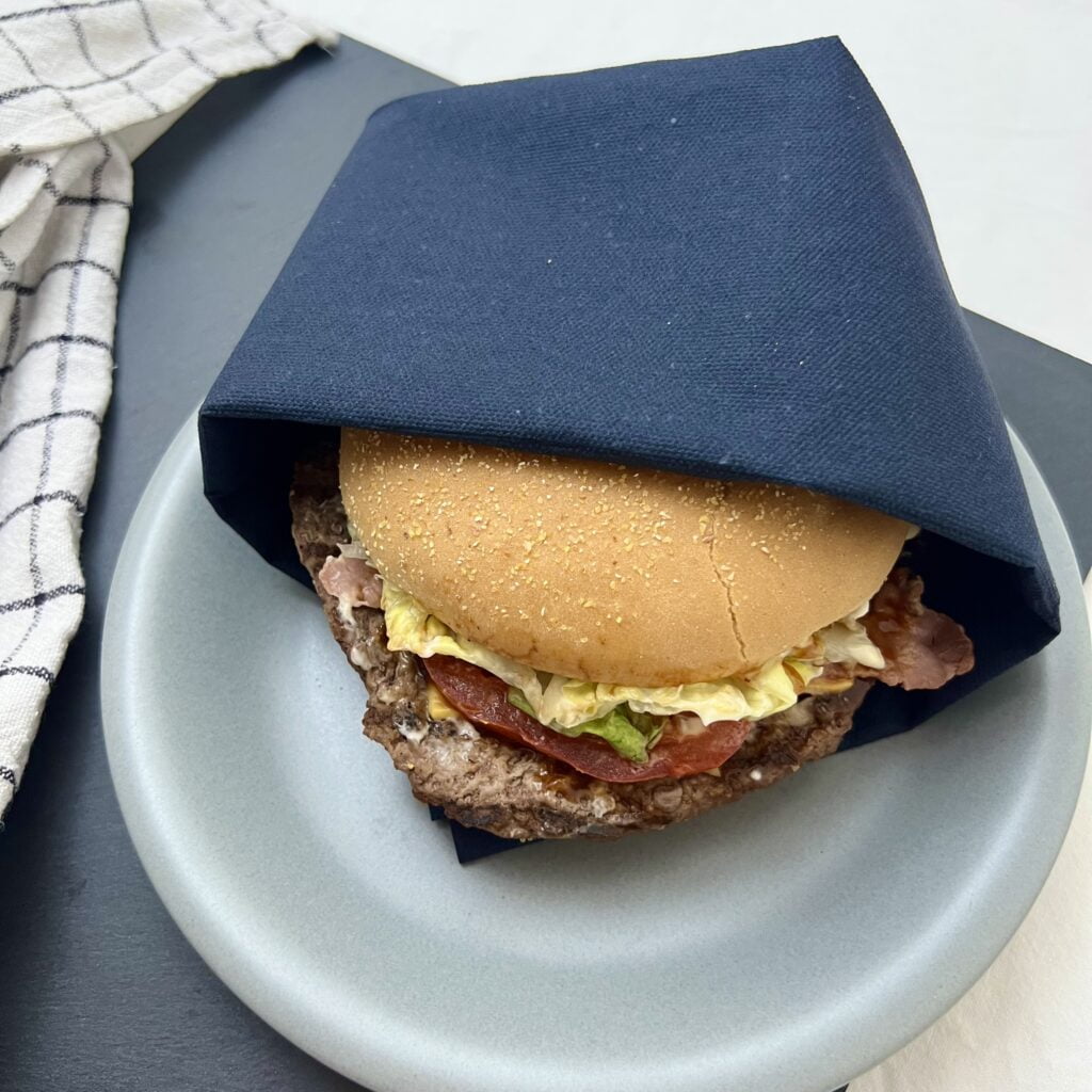 Hamburger Bulette im Burger bunt auf grauem teller in dunkler serviette auf dunklem untergrund neben weissem tuch
