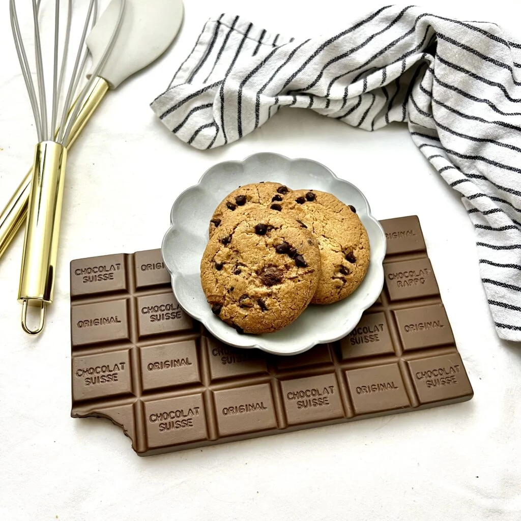 Schokoladen Cookies auf braunem schokoladen untergrund auf grauem teller mit weissem gestreiftem tuch im hintergrund auf weissem untergrund neben goldenen schwingbesen