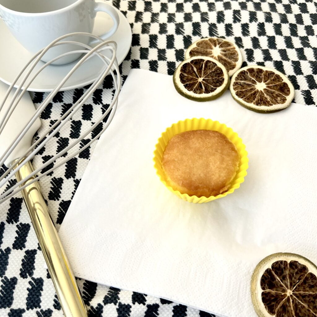 Zitronen Kokos Muffin goldbraun auf weisser Serviette neben Zitronenscheiben auf karriertem tuch schwarz weiss neben weisser tasse neben schwingbesen