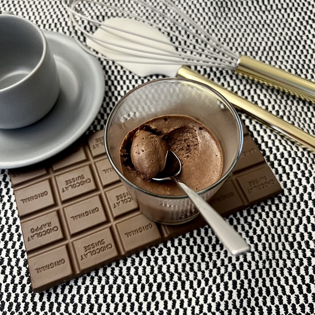 Schnelles braunes Schokoladenmousse auf brauner Schokoladentafel auf kariertem untergund neben grauen tasse neben schwingbesen