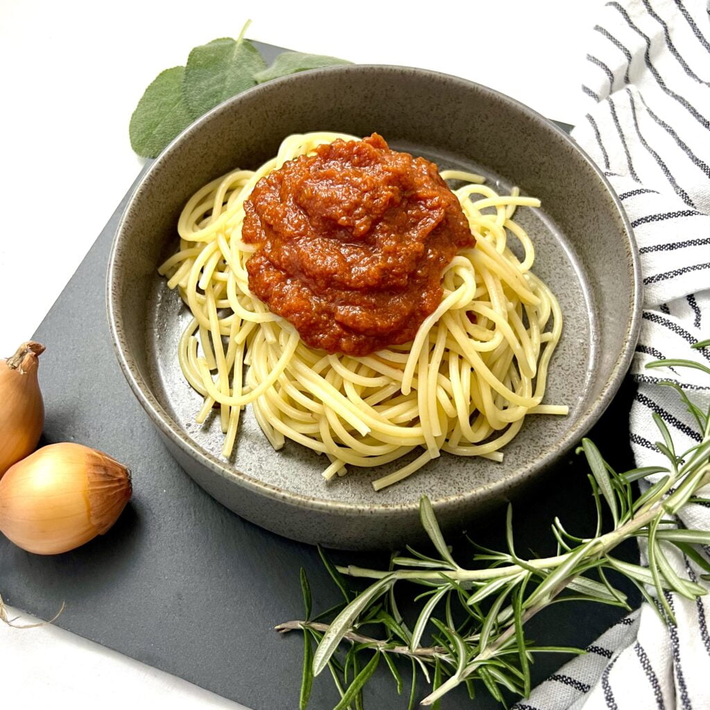 sugo rezept italienisch traditionell tomatensauce rot neben gelben spaghetti auf grauem teller auf dunklem untergrund mit zwiebeln und thymian im vordergrund